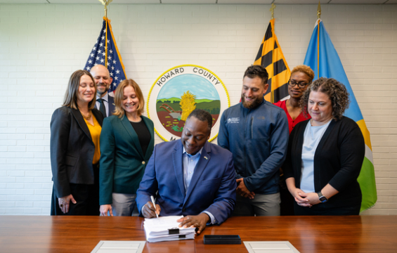 Howard County Executive Calvin Ball signs a document as six people stand behind him beneath the county seal.