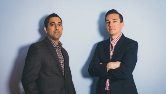 Two men in dark blazers and checked shirts stand against a light blue wall.