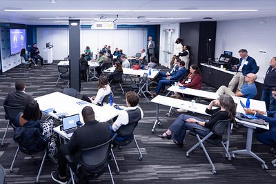 Attendees sit at tables with laptops and notebooks facing a presenter and projection screens in a meeting room.